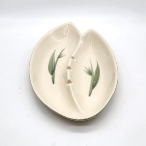 May include: Cream-coloured ceramic ashtray in a leaf-like shape. The ashtray has two sections and a central rest area. It is decorated with green floral designs and a green trim.