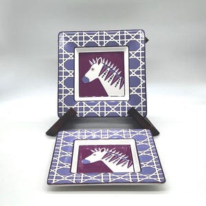 May include: Two square decorative plates with a zebra design. The plates have a purple and white geometric border and a central square with a zebra illustration in white, purple, and black. The plates are displayed on small brown stands.