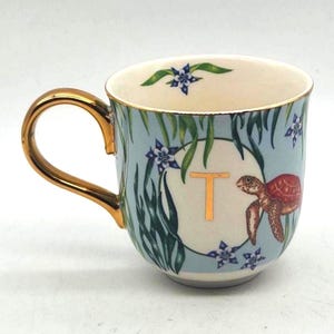 Puede incluir: Una taza de cerámica con asa y borde dorados. La taza presenta un fondo azul claro con un diseño de algas, flores y una tortuga. La letra "T" es dorada. La taza es un artículo decorativo.