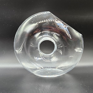 May include: A clear glass bowl with a circular opening in the center. The bowl has a curved, asymmetrical shape with a slightly raised rim. The glass is transparent, allowing light to pass through.