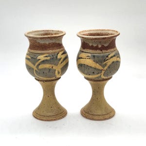 Op de afbeelding: Twee handgemaakte keramische kelken. Elke kelk heeft een beige voet en steel, een grijze body met gele abstracte ontwerpen en een bruine rand. De kelken worden gepresenteerd tegen een witte achtergrond.