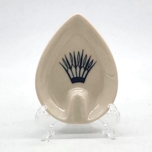 May include: A beige, teardrop-shaped ashtray with a dark blue, stylized crown design. The ashtray is made of ceramic and sits on a clear, plastic stand. The design is centered on the ashtray.