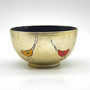 May include: A gold-colored bowl with a black interior. The bowl features two stylized bird illustrations, one yellow and the other red. The birds have black outlines and are positioned on the bowl's exterior.