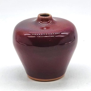 May include: A small, bulbous ceramic vase with a deep, glossy oxblood red glaze. The vessel features a narrow, short neck and a rounded body that tapers toward a flat, unglazed base showing the natural clay color. The surface has a smooth finish.