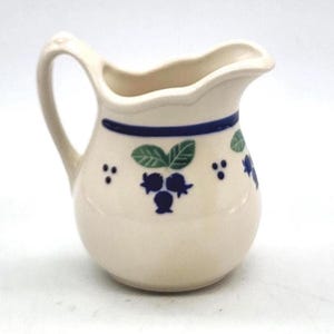 Pode incluir: Cremeira de cerâmica creme com uma alça curva e um bico. A cremeira é decorada com uma faixa azul na parte superior e desenhos de mirtilos e folhas azuis. A cremeira tem aproximadamente 10 cm de altura.