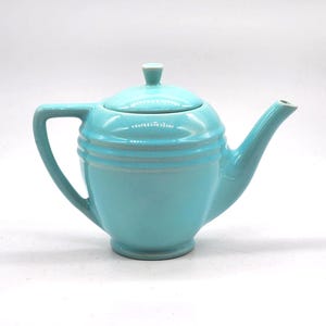 May include: A turquoise ceramic teapot with a lid and a curved handle. The teapot has a spout and a rounded body with horizontal ridges. The handle is a curved, open design. The teapot is set against a white background.