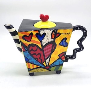 Puede incluir: Una tetera de cerámica con forma triangular, que presenta una tapa amarilla rematada con un corazón rojo. El cuerpo muestra patrones geométricos, corazones y rayas vibrantes y multicolores. Tiene un pico a rayas blancas y negras y un asa negra ondulada.