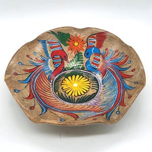 May include: Hand-painted, decorative ceramic bowl with a scalloped edge. The bowl features a central floral design and two stylized birds in red, blue, and yellow. The background is a tan color.