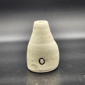 Puede incluir: Pequeño jarrón de cerámica blanquecina, cuerpo redondeado y cuello estrecho. Superficie texturizada, detalle de círculo negro y pequeño agujero. Probablemente hecho a mano, ideal para pequeños arreglos florales.