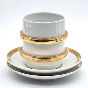 Könnte beinhalten: Ein weißes Tee-Set mit goldenen Verzierungen. Die Tasse hat eine zylindrische Form mit zwei goldenen Bändern. Die Untertassen sind gestapelt und haben ebenfalls goldene Ränder. Das Set befindet sich auf weißem Hintergrund.