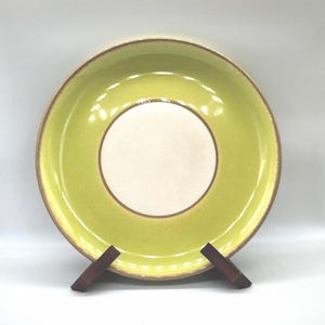 May include: A round, ceramic plate with a light green exterior and a cream-coloured centre. The plate has a brown rim and is displayed on a wooden stand. The plate is a decorative item.