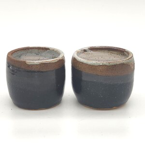 Puede incluir: Dos pequeñas tazas de cerámica hechas a mano. Las tazas son de color azul oscuro con una banda marrón alrededor de la parte superior y un interior más claro. Las tazas son redondas y tienen un aspecto rústico y texturizado.