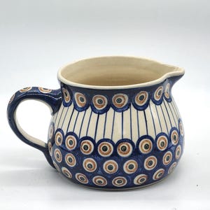 Może przedstawiać: Ceramiczny dzbanek o zaokrąglonym korpusie, uchwycie i wylewce. Dzbanek jest zdobiony niebiesko-białym wzorem z brązowymi i niebieskimi okręgami. Wnętrze jest jasno beżowe.