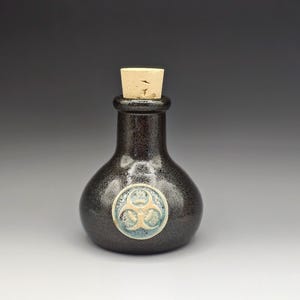 May include: A dark, speckled ceramic bottle with a cork stopper. The bottle has a round body and a narrow neck. A circular emblem with a biohazard symbol is affixed to the front. The bottle is likely for decorative or display purposes.