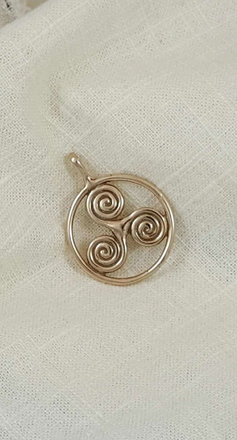 May include: A gold-toned pendant featuring a Celtic knot design with three intertwined spirals. The pendant is circular and has a loop at the top for attaching to a chain.