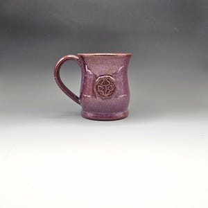 May include: A handmade ceramic mug in a deep purple hue, featuring a textured surface and a prominent handle. The mug showcases a raised emblem on its front. This unique mug is perfect for enjoying hot beverages.