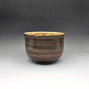 May include: A handmade ceramic cup with a dark brown exterior and a lighter brown interior. The cup has horizontal bands and a slightly textured surface. The rim is a lighter tan color.