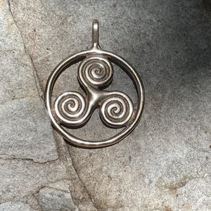 May include: A silver pendant featuring a Celtic knot design with three intertwined spirals. The pendant is circular and has a loop at the top for attaching to a chain.