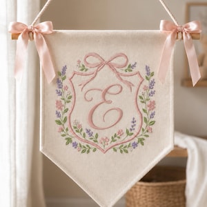 Puede incluir: Un estandarte de pared de tela color crema con una letra E rosa bordada en el centro de una corona floral de tallos lavanda, rosas y verdes. El estandarte está suspendido por una cuerda y adornado con dos lazos de satén rosa claro en las esquinas superiores.