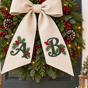 May include: A holiday wreath made of green pine branches, pinecones, and red berries, illuminated by small warm lights. A large beige linen bow hangs from the top, featuring the embroidered green letters A and B, each accented with small red berries.
