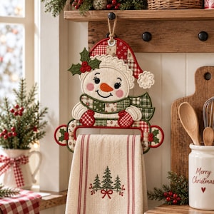 May include: A fabric snowman towel holder featuring a red and white plaid hat, a green and white plaid scarf, and embroidered holly leaves. A cream-colored kitchen towel with red stripes and an embroidered pine tree design hangs from the snowman.