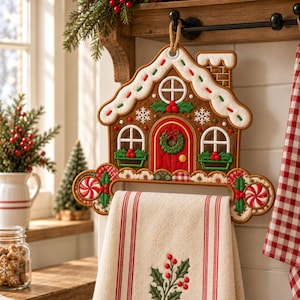May include: A gingerbread house-shaped towel holder featuring a red door, white icing trim, green holly accents, and peppermint candy details. The holder is made of fabric and includes a beige kitchen towel with red stripes and a holly embroidery design.