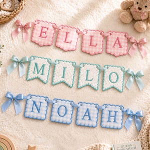 May include: Three fabric banners featuring embroidered names. The top banner is pink gingham with the name ELLA. The middle banner is white with teal polka dots and the name MILO. The bottom banner is blue gingham with the name NOAH. Each banner has satin bows.