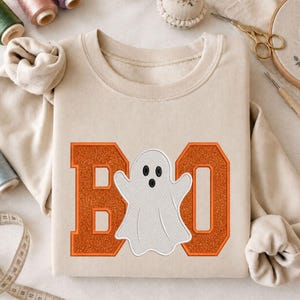 May include: A beige crewneck sweatshirt featuring a large graphic on the front. The design consists of two orange glittery letters, B and O, with a white embroidered ghost positioned in the center to spell the word BOO.