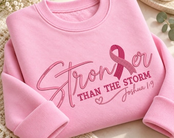May include: A light pink long-sleeve sweatshirt featuring the embroidered text "Stronger than the storm" with a pink awareness ribbon replacing the letter "g" and the scripture reference "Joshua 1:9" written below with a small heart icon.