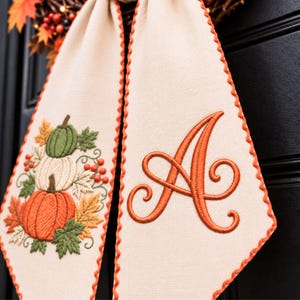 May include: A cream-colored fabric ribbon with scalloped orange edges. One side features an embroidered stack of green, white, and orange pumpkins with autumn leaves and berries. The other side displays a large, embroidered orange letter A.