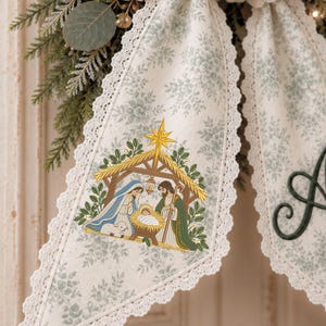 May include: A cream-colored fabric ribbon with a delicate sage green floral pattern and white lace trim edges. The ribbon features a detailed, colorful embroidered nativity scene depicting a stable, a star, and three figures.