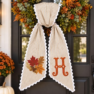 May include: A beige and white striped fabric door hanger featuring a scalloped white lace trim. The left panel displays an embroidered autumn leaf and acorn motif, while the right panel features a large, decorative orange embroidered letter H.