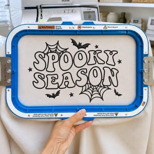 May include: A rectangular blue embroidery hoop holding off-white fabric with black machine-embroidered text that reads SPOOKY SEASON. The design includes black spiderwebs, small stars, and bat silhouettes. Warning labels are visible on the frame.