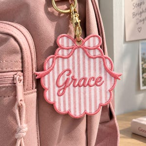 May include: A pink and white striped fabric bag charm featuring a scalloped edge and a bow design at the top. The name Grace is embroidered in pink script across the centre. The charm is attached to a gold-toned metal lobster clasp and ring.