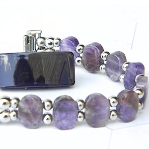 Puede incluir: Una pulsera con piedras preciosas ovaladas de color p&uacute;rpura y peque&ntilde;as cuentas plateadas. La pulsera tiene un cierre plateado y un elemento rectangular azul oscuro. El fondo es blanco.