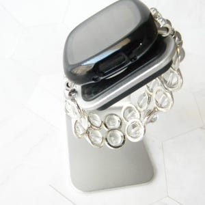 May include: A black smartwatch with a silver band is displayed on a silver stand. The band is made of clear, round beads.