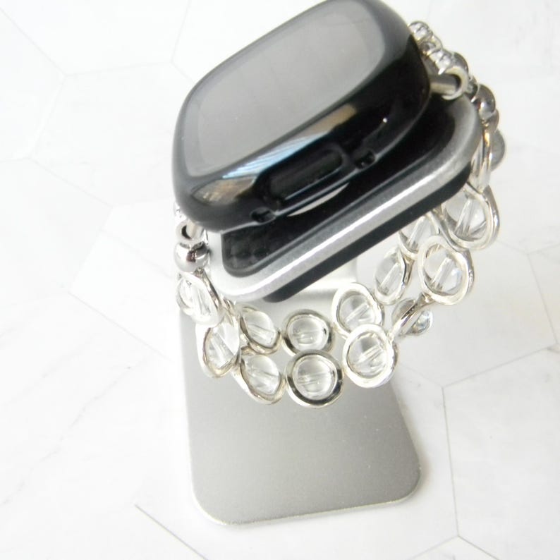 May include: A black smartwatch with a silver band is displayed on a silver stand. The band is made of clear, round beads.