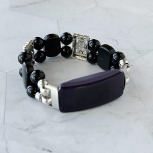 May include: A beaded bracelet featuring a dark purple rectangular center piece and black, rounded beads. The bracelet has silver-toned metal accents and a clasp. The jewelry is displayed on a white and gray surface.