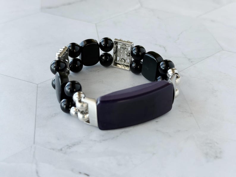 May include: A beaded bracelet featuring a dark purple rectangular center piece and black, rounded beads. The bracelet has silver-toned metal accents and a clasp. The jewelry is displayed on a white and gray surface.