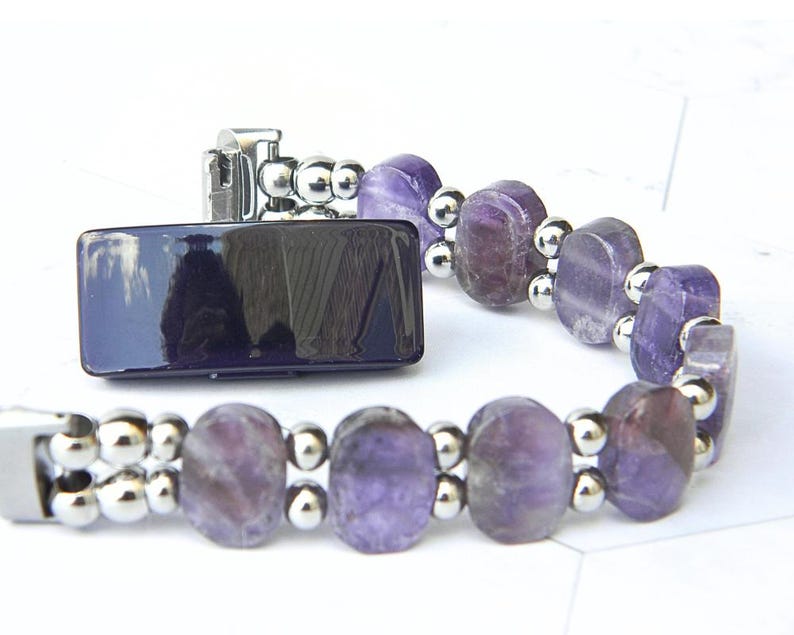 Puede incluir: Una pulsera con piedras preciosas ovaladas de color p&uacute;rpura y peque&ntilde;as cuentas plateadas. La pulsera tiene un cierre plateado y un elemento rectangular azul oscuro. El fondo es blanco.