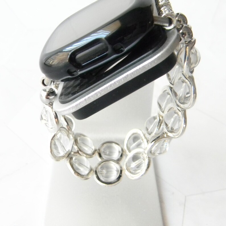 May include: A silver watch band with clear crystal beads. The watch band is designed to fit a square-shaped smartwatch.