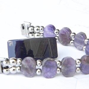 Puede incluir: Un brazalete con piedras preciosas ovales de color p&uacute;rpura y peque&ntilde;as cuentas plateadas. El brazalete tiene un cierre plateado y se muestra junto a un objeto rectangular azul oscuro. Las piedras preciosas tienen tonos p&uacute;rpuras claros y oscuros.