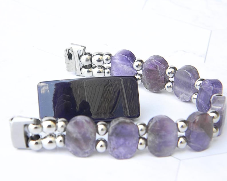 Puede incluir: Un brazalete con piedras preciosas ovales de color p&uacute;rpura y peque&ntilde;as cuentas plateadas. El brazalete tiene un cierre plateado y se muestra junto a un objeto rectangular azul oscuro. Las piedras preciosas tienen tonos p&uacute;rpuras claros y oscuros.