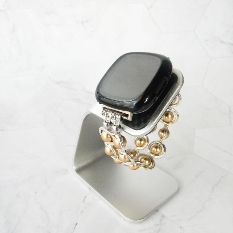 May include: A black smartwatch with a gold and silver beaded band on a silver stand.