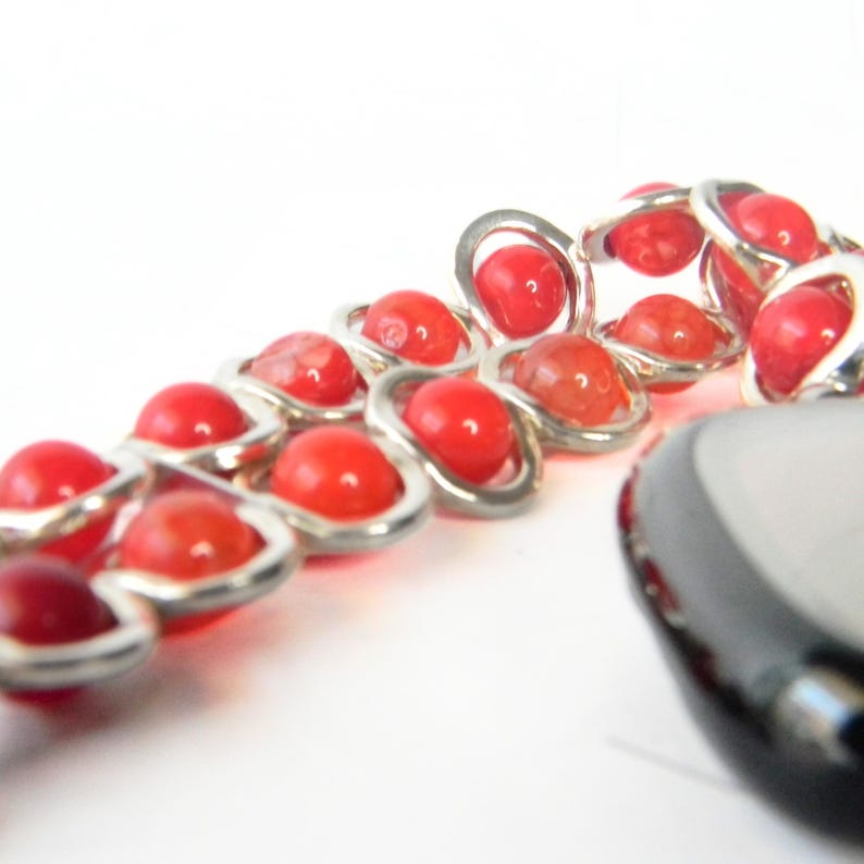 May include: A silver chain bracelet with red beads. The bracelet is made of small, interlocking silver rings with red beads in each ring. The bracelet is accented with a black stone pendant.
