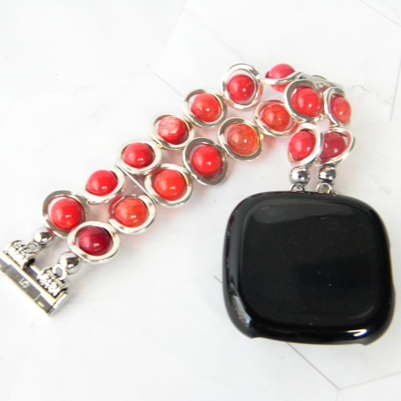 May include: A red and orange beaded watch band with a silver clasp. The band is made of two rows of beads, and the beads are graduated in size. The band is attached to a black watch face.