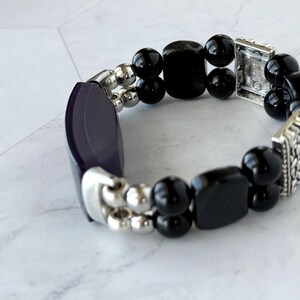 May include: A stretch bracelet featuring a combination of black, rounded beads and rectangular beads. The bracelet also includes silver-toned metal accents and a dark purple, curved rectangular piece.