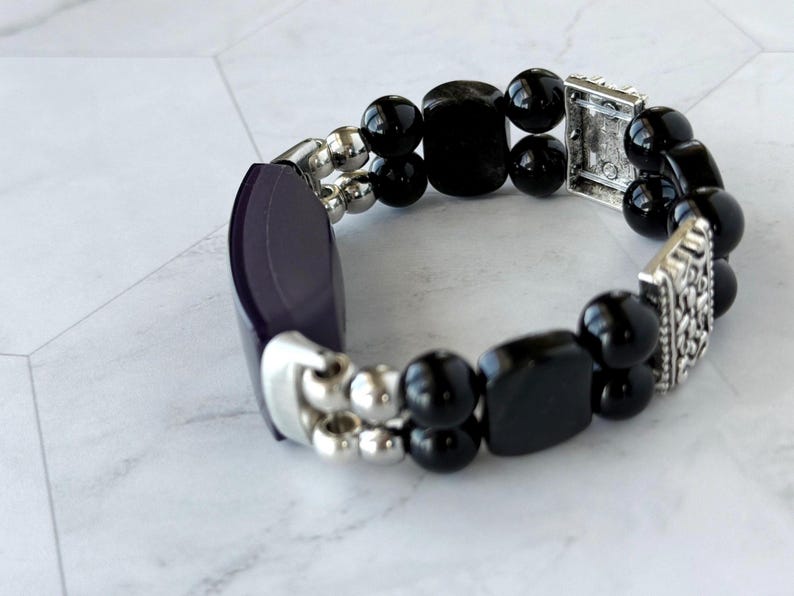 May include: A stretch bracelet featuring a combination of black, rounded beads and rectangular beads. The bracelet also includes silver-toned metal accents and a dark purple, curved rectangular piece.