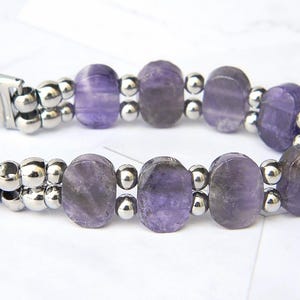 Puede incluir: Un brazalete con cuentas ovaladas de amatista p&uacute;rpura intercaladas con peque&ntilde;as cuentas met&aacute;licas plateadas. El brazalete tiene un cierre plateado y se muestra sobre una superficie blanca. Las cuentas de amatista tienen tonos p&uacute;rpuras claros y oscuros.