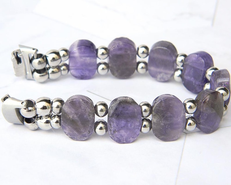 Puede incluir: Un brazalete con cuentas ovaladas de amatista p&uacute;rpura intercaladas con peque&ntilde;as cuentas met&aacute;licas plateadas. El brazalete tiene un cierre plateado y se muestra sobre una superficie blanca. Las cuentas de amatista tienen tonos p&uacute;rpuras claros y oscuros.